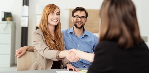 Happy young couple meeting with a broker in her office leaning over the desk to shake hands, view from behind the female agent-1